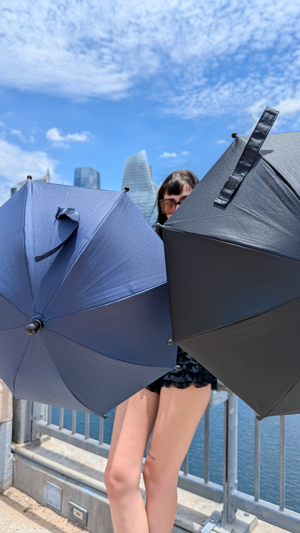 A woman wearing sunglasses stands behind two large umbrellas, one dark blue and one black, with a city skyline and water in the background under a blue sky.