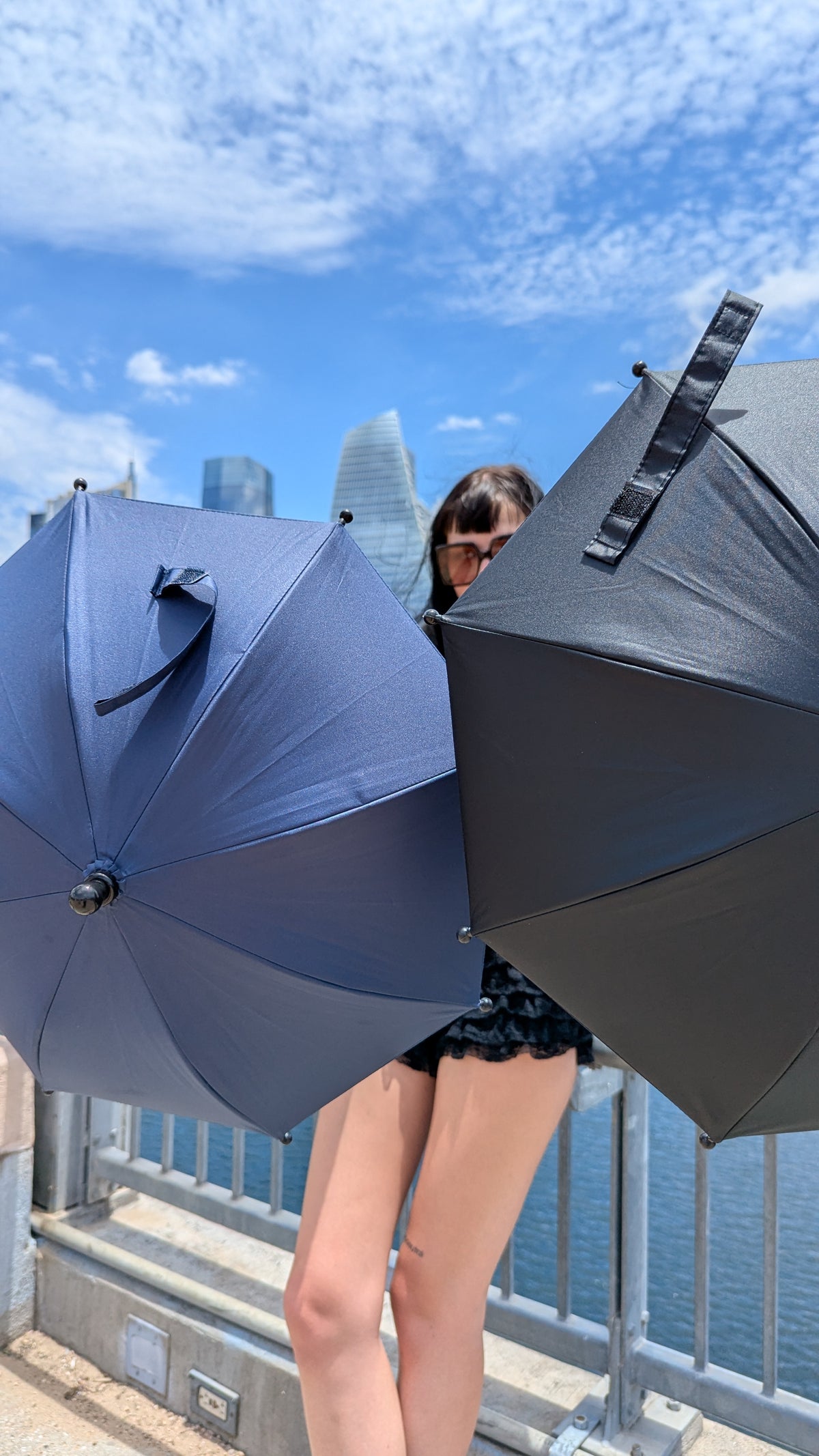 A woman wearing sunglasses stands behind two large umbrellas, one dark blue and one black, with a city skyline and water in the background under a blue sky.