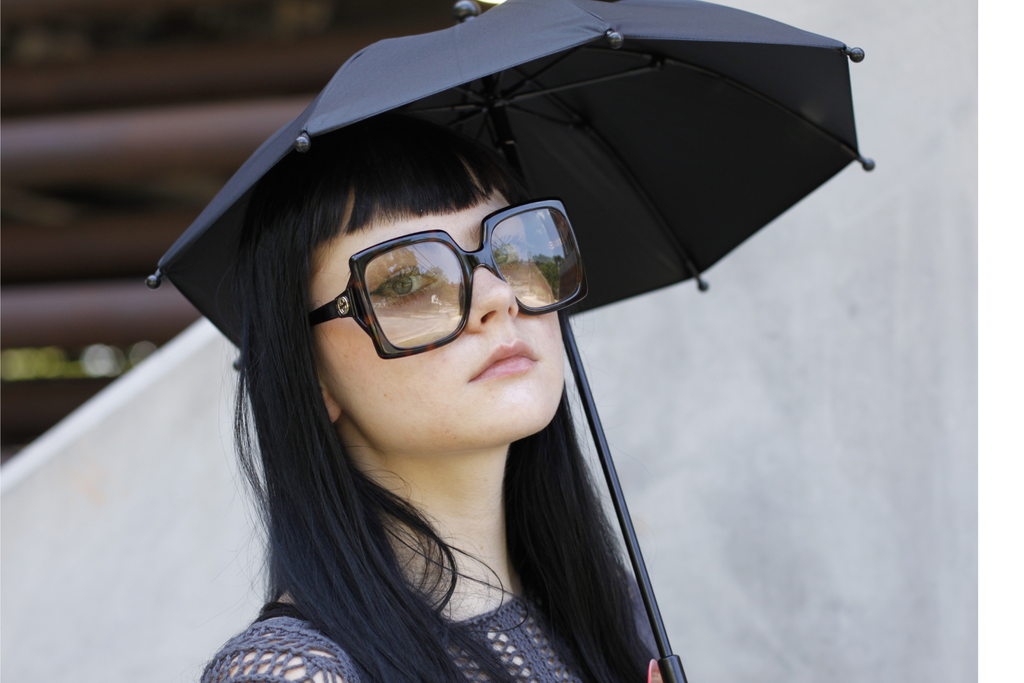 A woman with long black hair wearing oversized square sunglasses and holding a black Stroll Shade Parasol, photographed against a light gray wall.