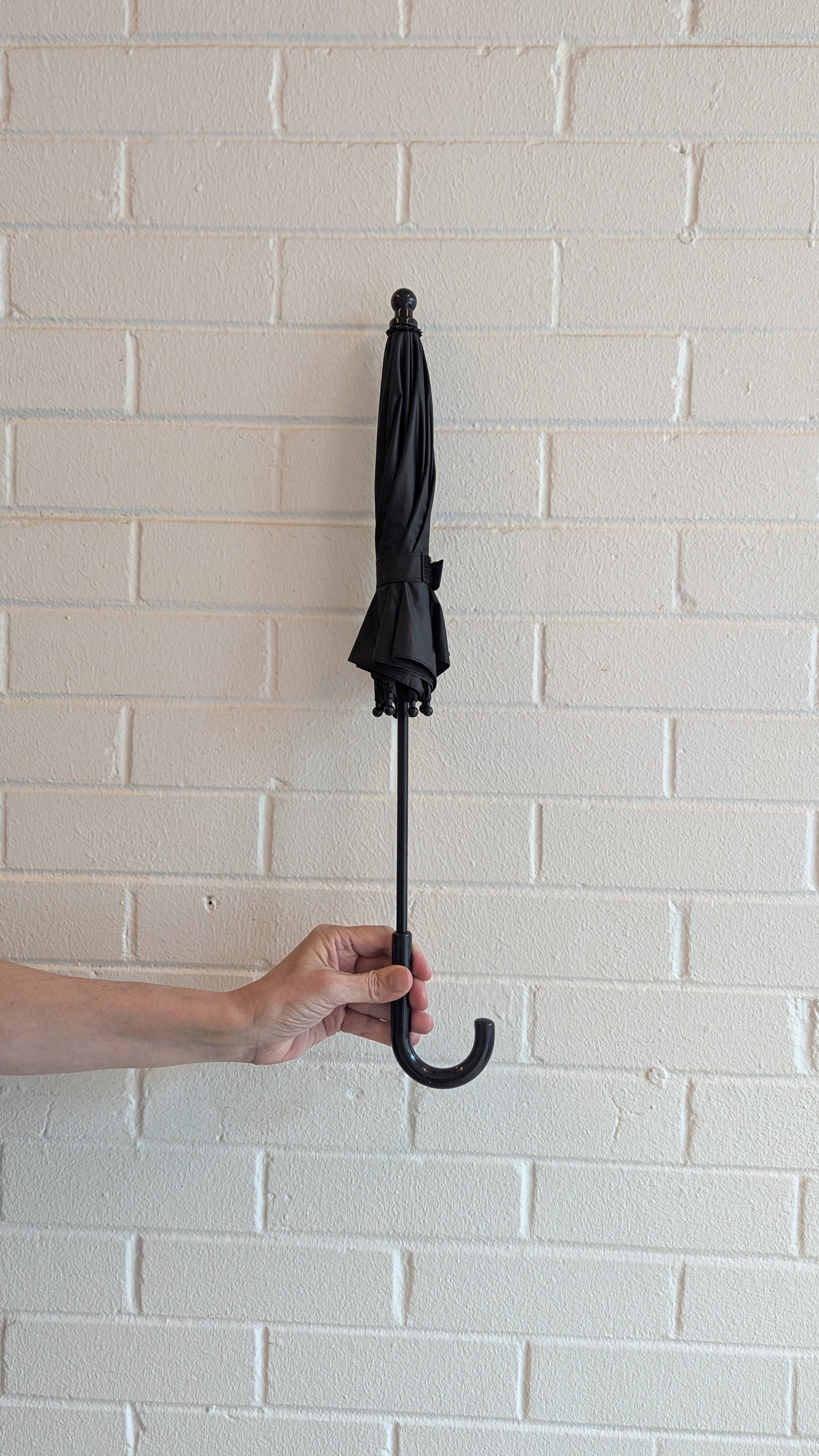 Person holding a black parasol closed against a white brick wall